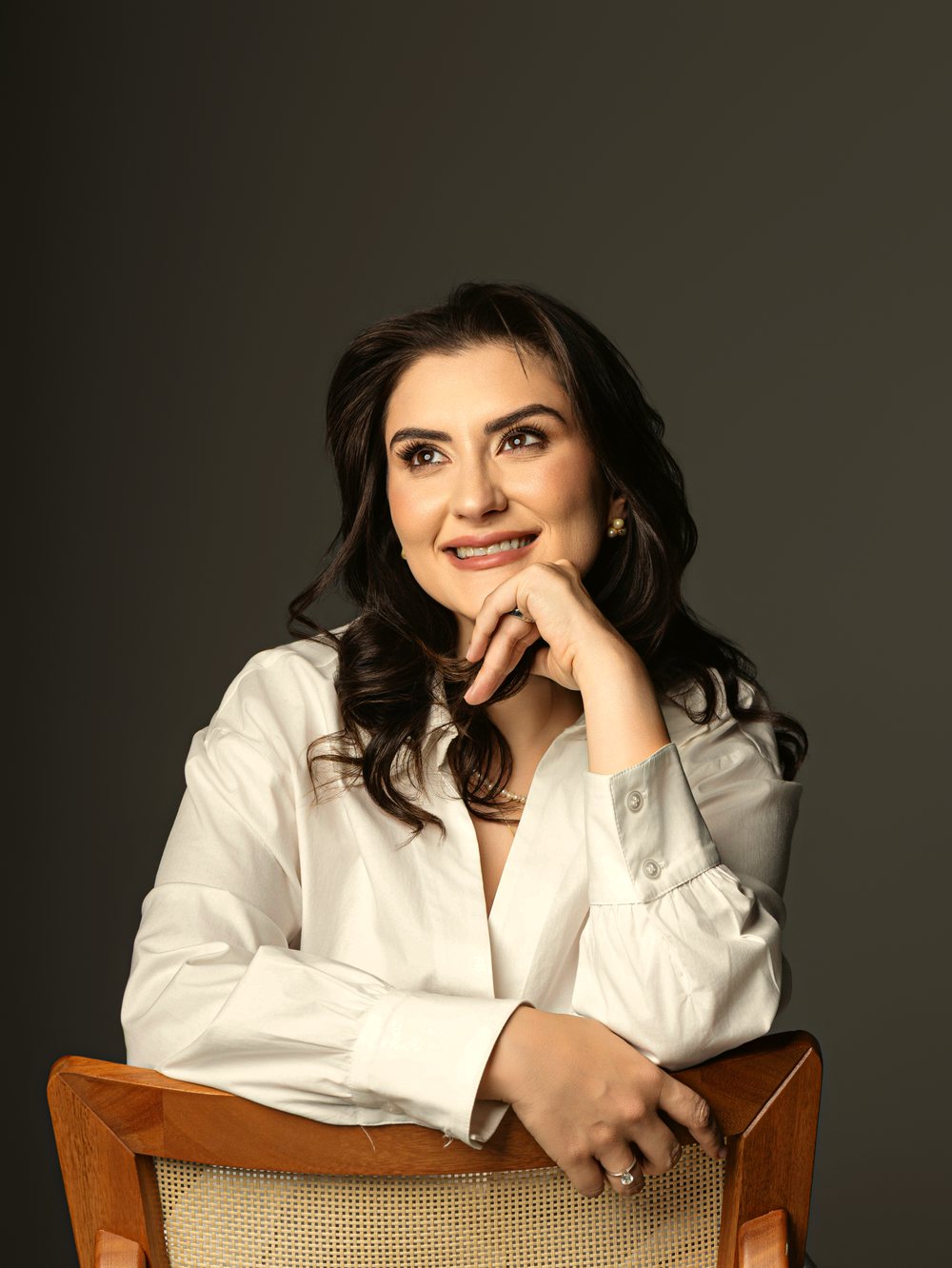 Dra. Marcella Pagnano Bocchi sorrindo, apoiada em uma cadeira de madeira, em um retrato de luz natural